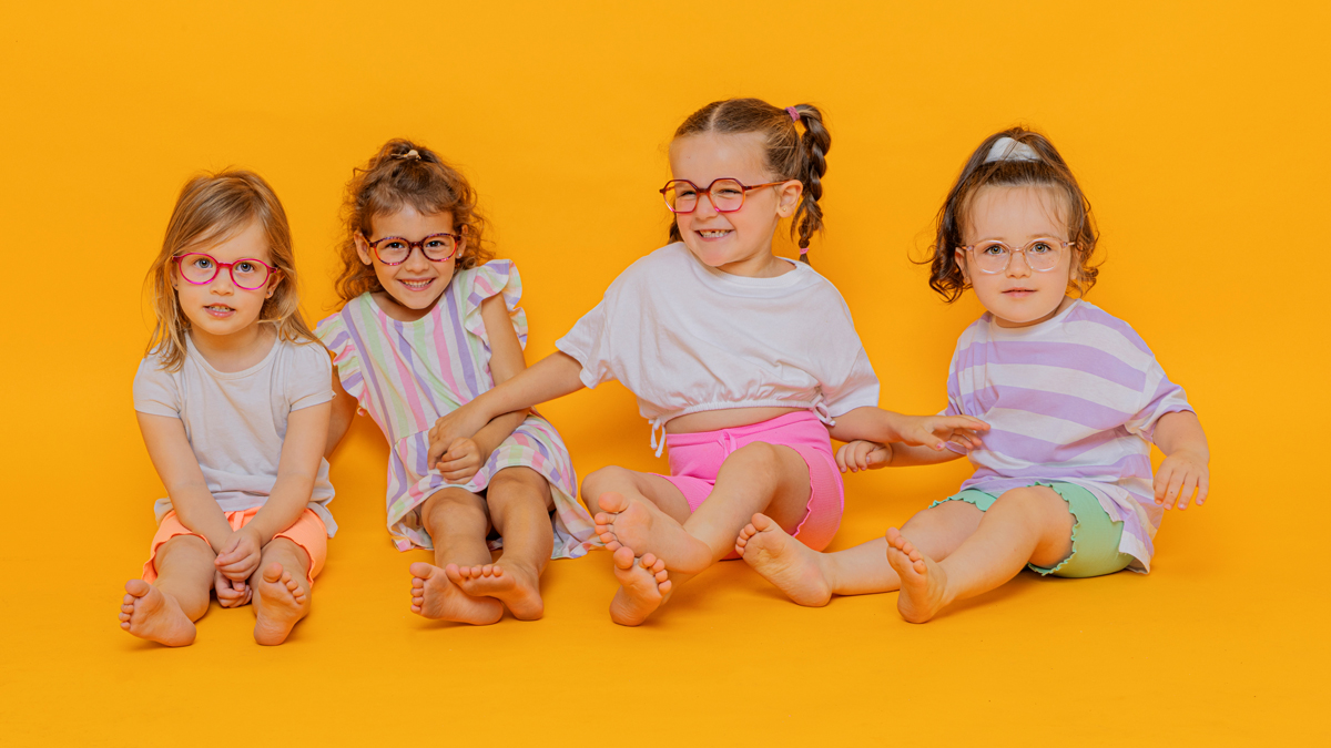 Quatre petites filles souriantes, assises par terre, les mains sur les genoux, chacune portant des lunettes